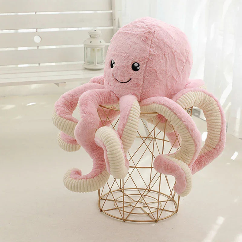 Peluche pieuvre rose souriante avec tentacules beige, posée sur un fauteuil design en fil métallique