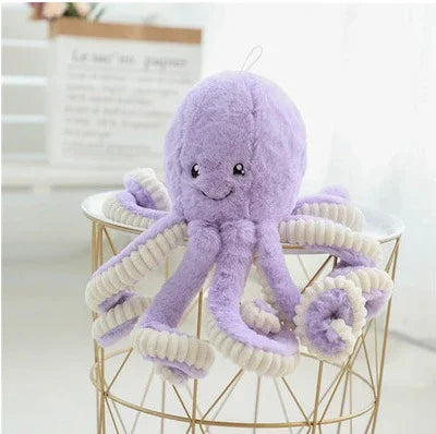 Peluche pieuvre violette avec tentacules torsadés crème, posée sur un panier métallique doré