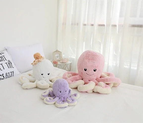 Peluches pieuvres colorées rose, violet et blanc sur un lit blanc près d'une fenêtre avec rideaux