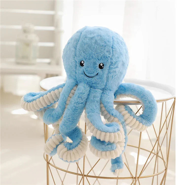 Peluche pieuvre bleue douce avec visage souriant, pose sur une table dorée dans une pièce lumineuse