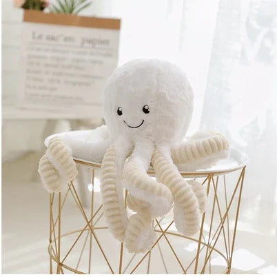 Peluche pieuvre blanche souriante avec tentacules rembourrées, posée sur table moderne dorée dans pièce lumineuse